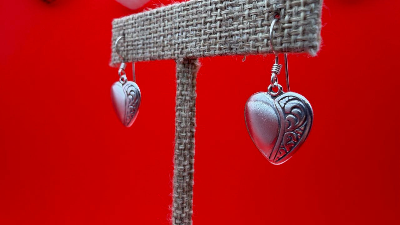 Heart-shaped silver earrings on a burlap stand against a red background