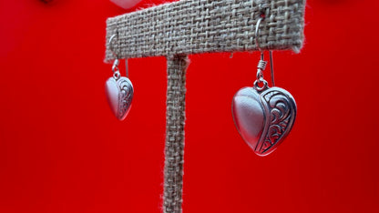 Heart-shaped silver earrings on a burlap stand against a red background